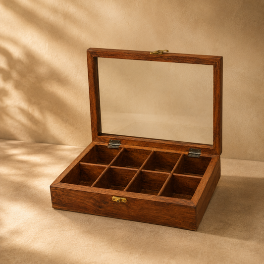 A rectangular wooden jewelry box with a hinged glass lid, shown open to reveal nine interior compartments. The box has a warm medium-brown wood finish with small brass-tone metal latches and hinges, set against a beige textured backdrop with soft shadow overlays.