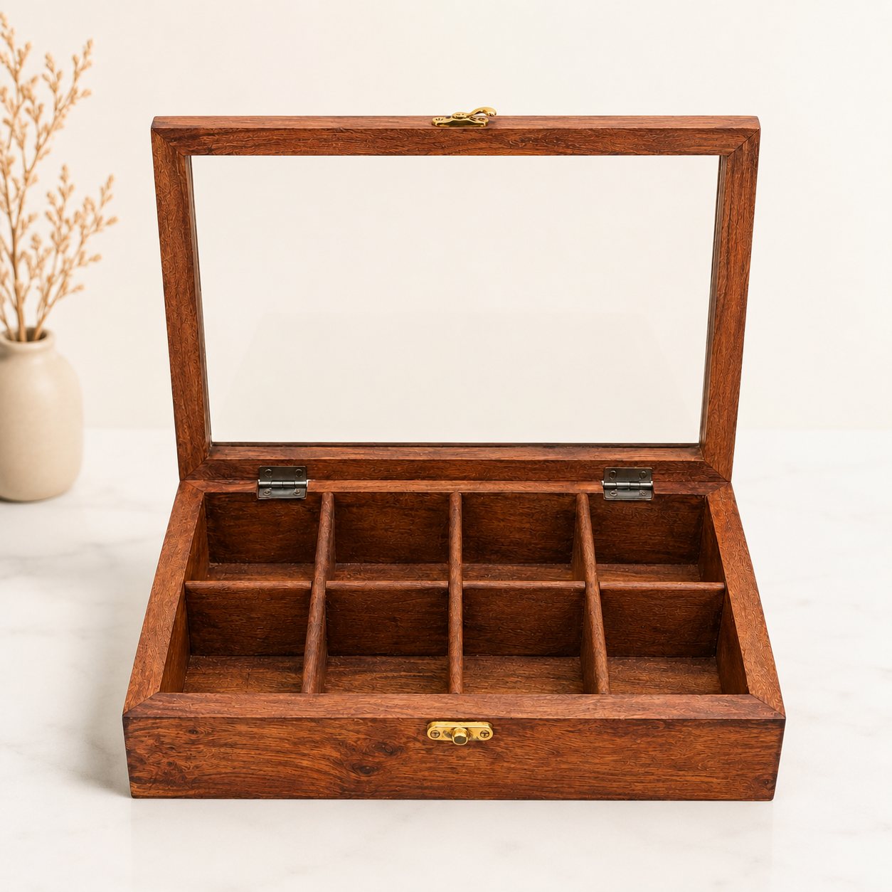 A wooden jewelry organizer box with a glass lid, shown open to reveal nine compartments. The box has a warm medium-brown wood finish with small brass-tone metal accents on the latch and hinge, set against a clean white background with a small vase of dried stems in the background.