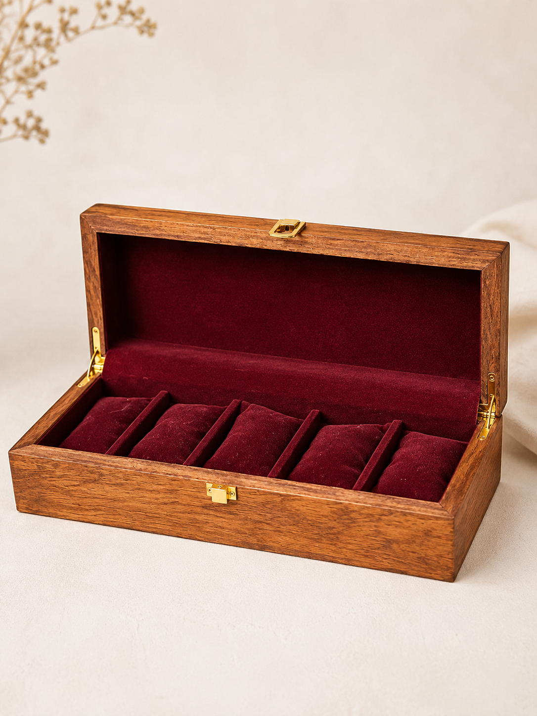 An open wooden jewelry box with a dark red velvet interior and five ring slots, shown on a light beige background.